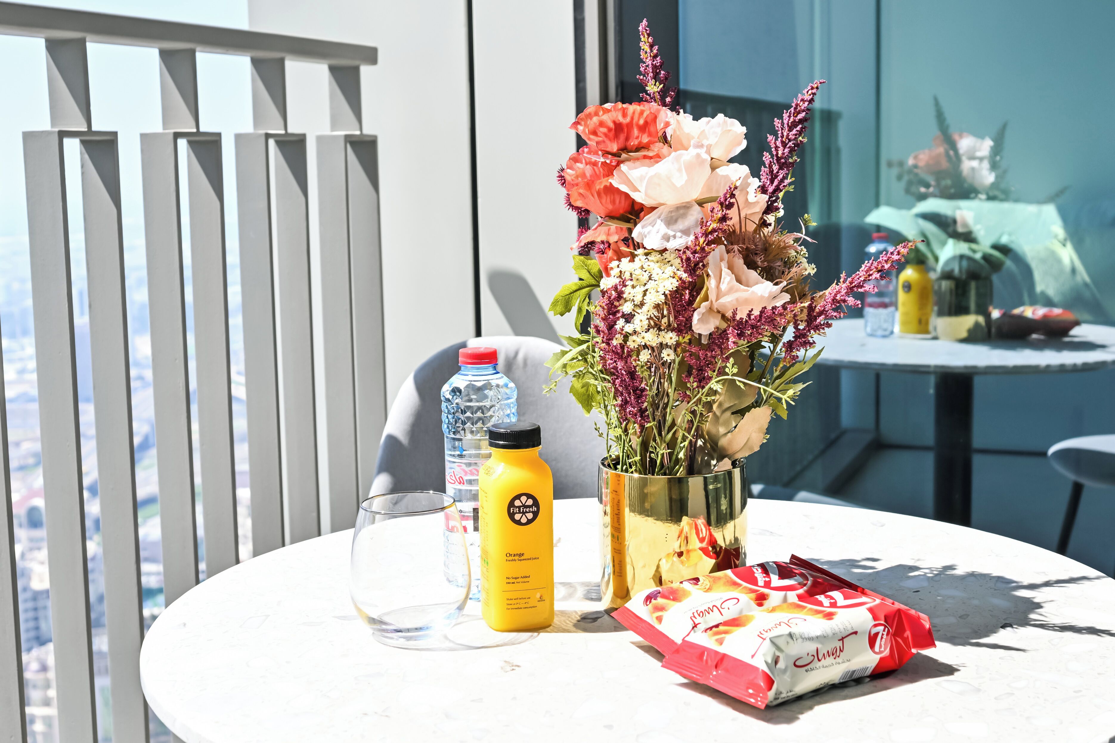 A table with a glass, a bottle, a vase with flowers, and a packet of snacks.