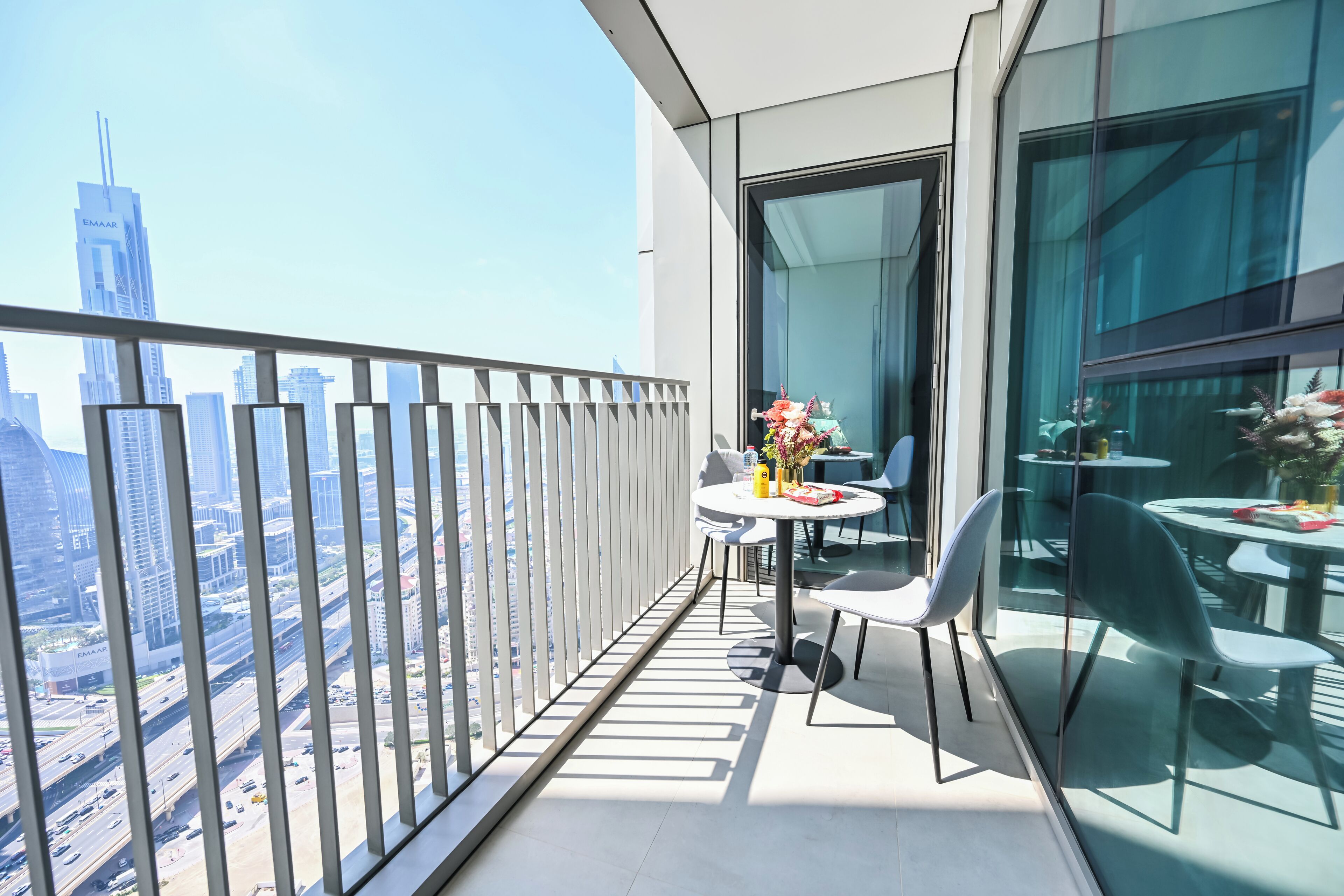 A balcony with a glass railing, a round table with chairs, and a vase with flowers.
