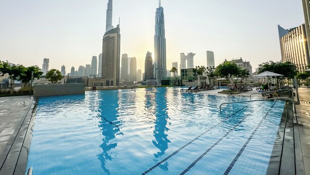 A swimming pool with clear blue water and lane dividers, surrounded by a wooden deck. In the background, there are high-rise buildings, including a prominent skyscraper. - Burj Khalifa View - 2 BR - PS5 - Pool (Dubai)