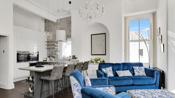 A modern kitchen with white cabinets, a central island, and bar stools. There is a blue sofa with patterned cushions and a chandelier above. - 2 Bed - Hoe Heights by Smart Welcomes (Plymouth)