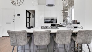 A modern kitchen with white cabinets, a central island with bar stools, a large clock, and pendant lights. - 2 Bed - Hoe Heights by Smart Welcomes (Plymouth)