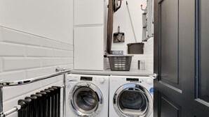 A compact laundry area with two front-loading washing machines, a drying rack, and a small ironing station. - 2 Bed - Hoe Heights by Smart Welcomes (Plymouth)