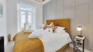 A neatly made bed with a yellow headboard, white bedding, and colorful pillows. There is a bedside table with a lamp and a small vase. - 2 Bed - Hoe Heights by Smart Welcomes (Plymouth)