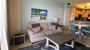 A living room with a beige sofa, a coffee table, and a painting of a beach scene. - SUNRISE BEACH 2105, 2 BED 2 BA. balcony hOt TUB PoolS! (panama city beach)