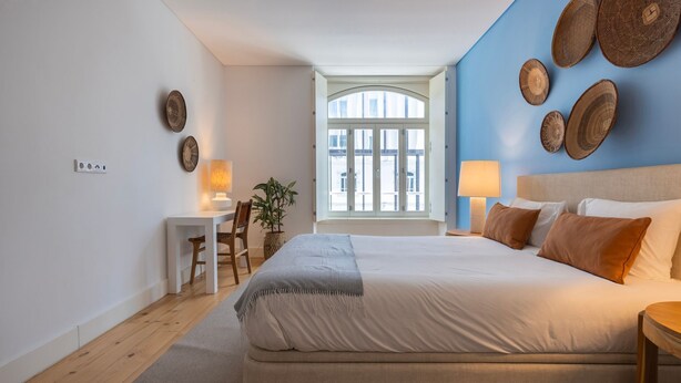A bedroom with a large bed, two bedside tables with lamps, a desk with a chair, and a view of the outside through a window.