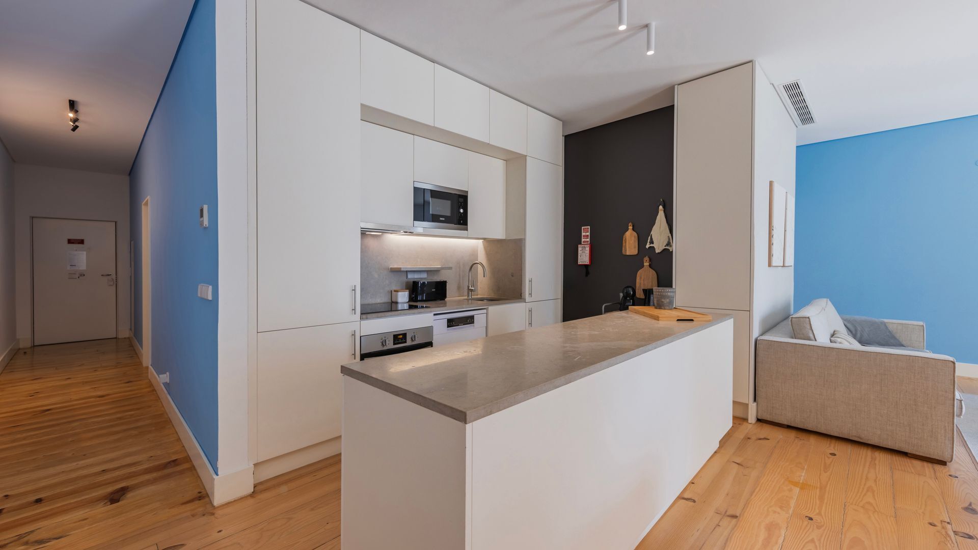 A modern kitchen with white cabinets, a microwave, and a built-in oven. There's a wooden floor and a blue accent wall.