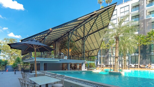 Zona de piscina al aire libre con área de descanso cubierta, palmeras y un edificio moderno al fondo.