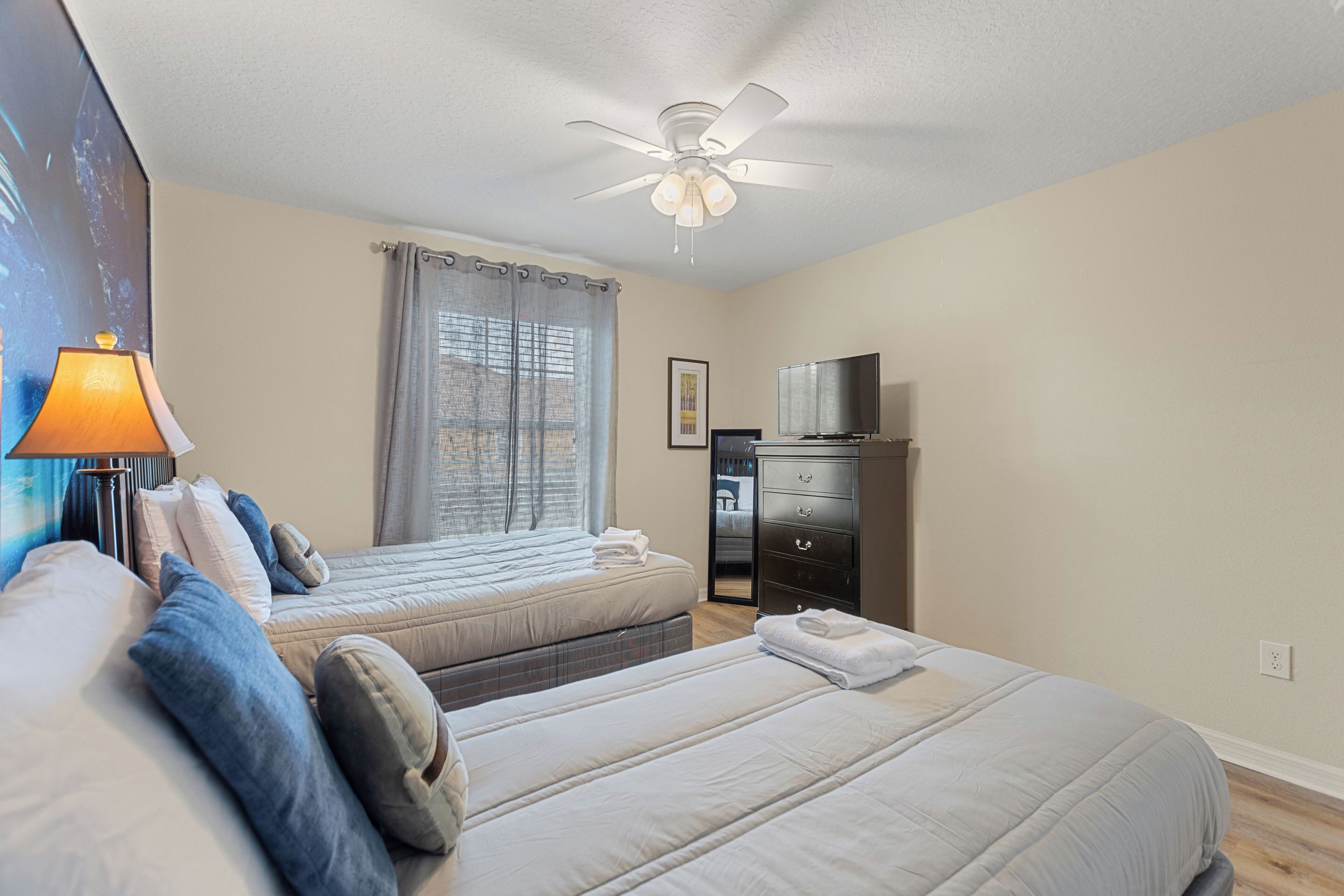 A hotel room with two beds, a ceiling fan, a TV, a desk, and a window with curtains.