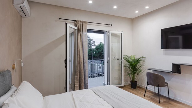 Une chambre moderne avec un lit, un téléviseur fixé au mur, une plante en pot et une porte-fenêtre coulissante donnant sur un balcon.