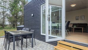 Une maison moderne dotée d’un patio comprenant une table et des chaises, un banc en bois et une porte-fenêtre coulissante donnant sur un espace intérieur avec un canapé et une table basse.