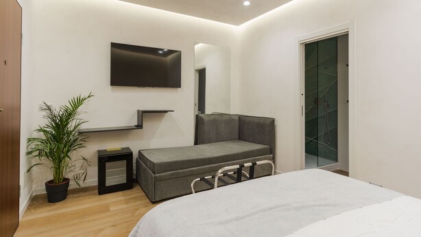Un dormitorio moderno con cama, un sofá gris, televisor montado en la pared, una planta en maceta y un vestidor.