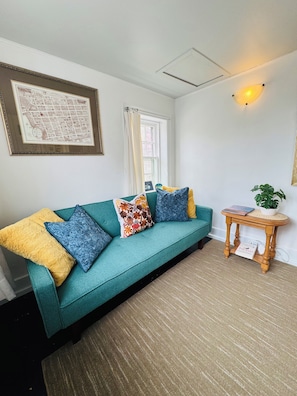 A teal sofa with patterned cushions, a framed map on the wall, a small wooden side table with a plant, and a window with curtains. - Heritage Cozy Historic home.  At the heart of downtown Charlottetown (Charlottetown)