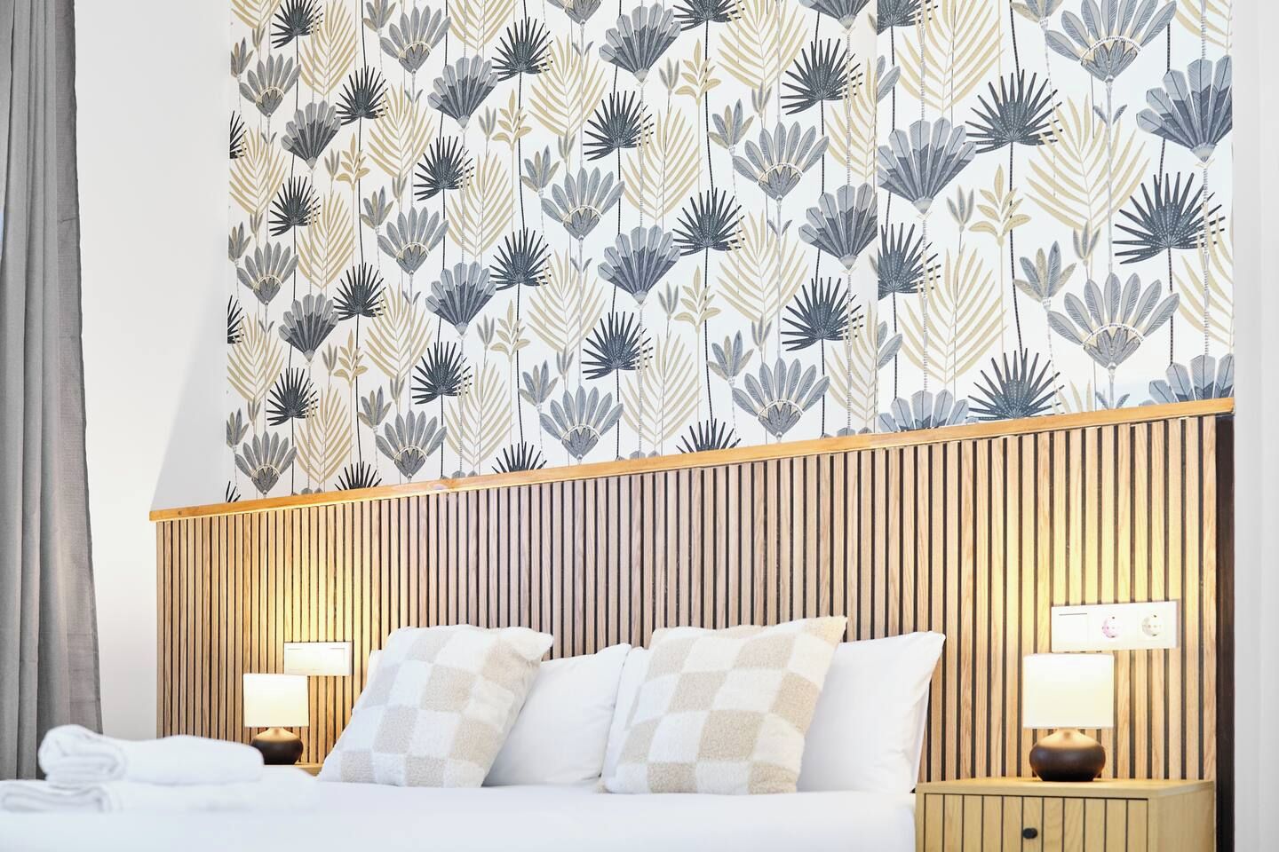 A hotel room with a bed, two bedside lamps, a wooden headboard, and a patterned wallpaper.