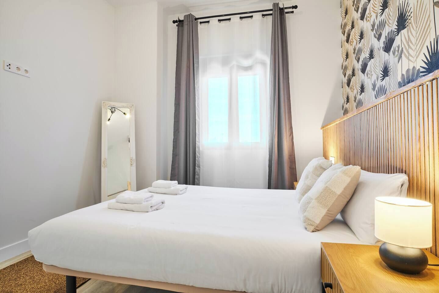 Habitación de hotel con una cama grande, una mesita de noche con lámpara, un espejo y una ventana con cortinas.