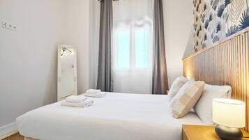 Habitación de hotel con una cama grande, una mesita de noche con lámpara, un espejo y una ventana con cortinas.