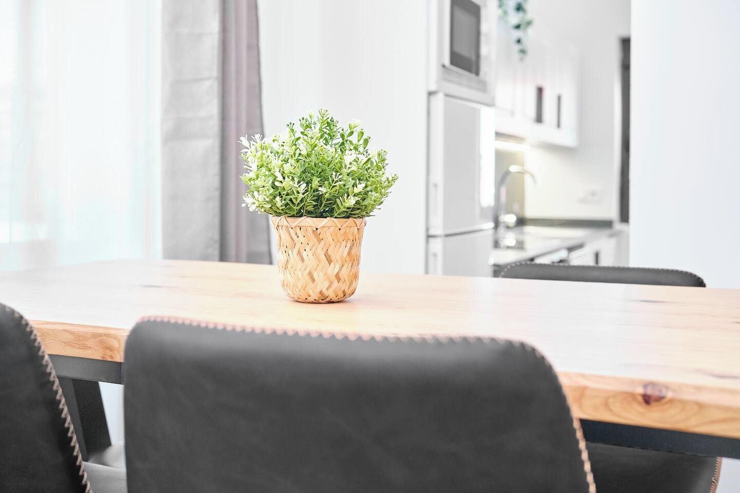 A dining table with a woven plant pot in the center, black chairs, and a kitchen in the background.