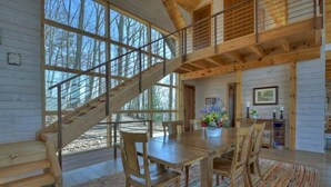 A wooden cabin interior with a dining area, staircase, and large windows.