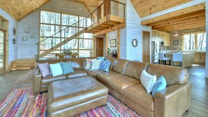 A spacious living area with a large sectional sofa, a wooden staircase, and a kitchen in the background.