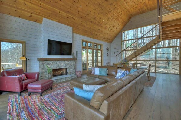 A spacious living room with a stone fireplace, leather sofas, and a large window overlooking a forest.