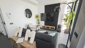 Ein modernes Wohnzimmer mit einem dunkelgrauen Sofa, einem Couchtisch mit Kissen, einem Flachbildfernseher und einer Stehlampe.