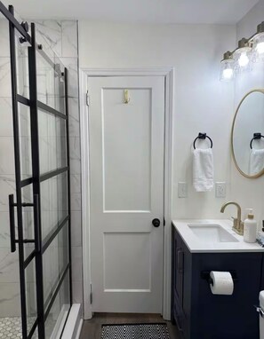 Ein Badezimmer mit einer Duschkabine aus Glas, einer weißen Tür, einem Waschtisch, einem Spiegel und einem Toilettenpapierhalter.
