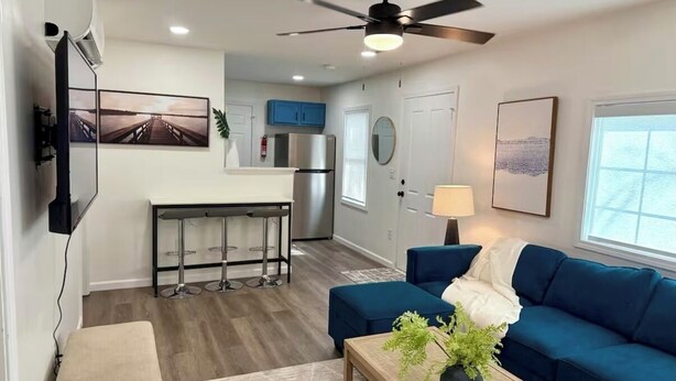 A modern living room with a blue sofa, a coffee table with a plant, and a kitchen area with a refrigerator and bar stools. - Cozy and Convenient- 5 min to Airport (North Charleston)