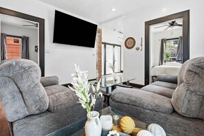 Ein modernes Wohnzimmer mit zwei grauen Sofas, einem Glastisch mit Dekoration, einem wandmontierten Fernseher und einem Deckenventilator.