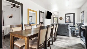 Un salon moderne avec une table à manger en bois, des chaises rembourrées de couleur beige, un canapé gris et un téléviseur à écran plat fixé au mur.