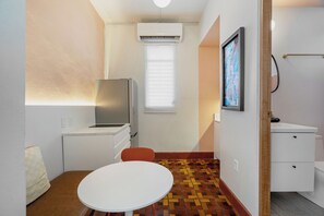 A compact room with a white table, a red chair, a small kitchenette, a window with blinds, and a framed picture on the wall.