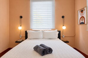 A hotel room with a bed, two bedside lamps, a window with blinds, a wall-mounted artwork, and a rolled towel on the bed.