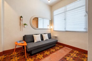 A living room with a grey sofa, a small orange side table, a mirror, and a window with blinds.