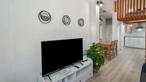 A modern living room with a flat-screen TV, a dining area with wooden chairs, and a kitchen in the background.
