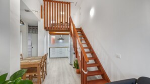 A staircase leading to an upper level, a dining area with a wooden table and chairs, and a kitchen area with white cabinets and a refrigerator.