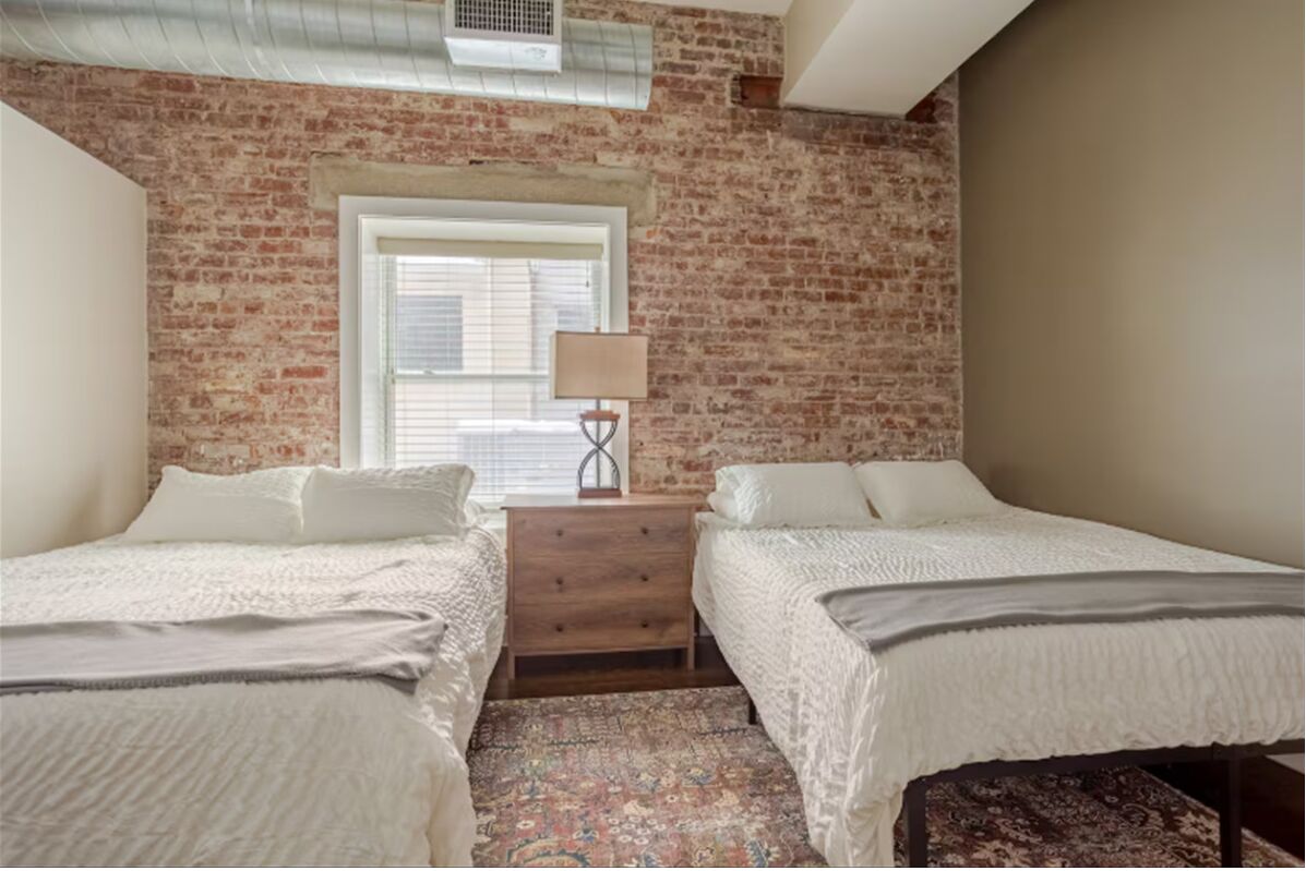 A bedroom with two beds, a wooden nightstand, a lamp, and a window with blinds.