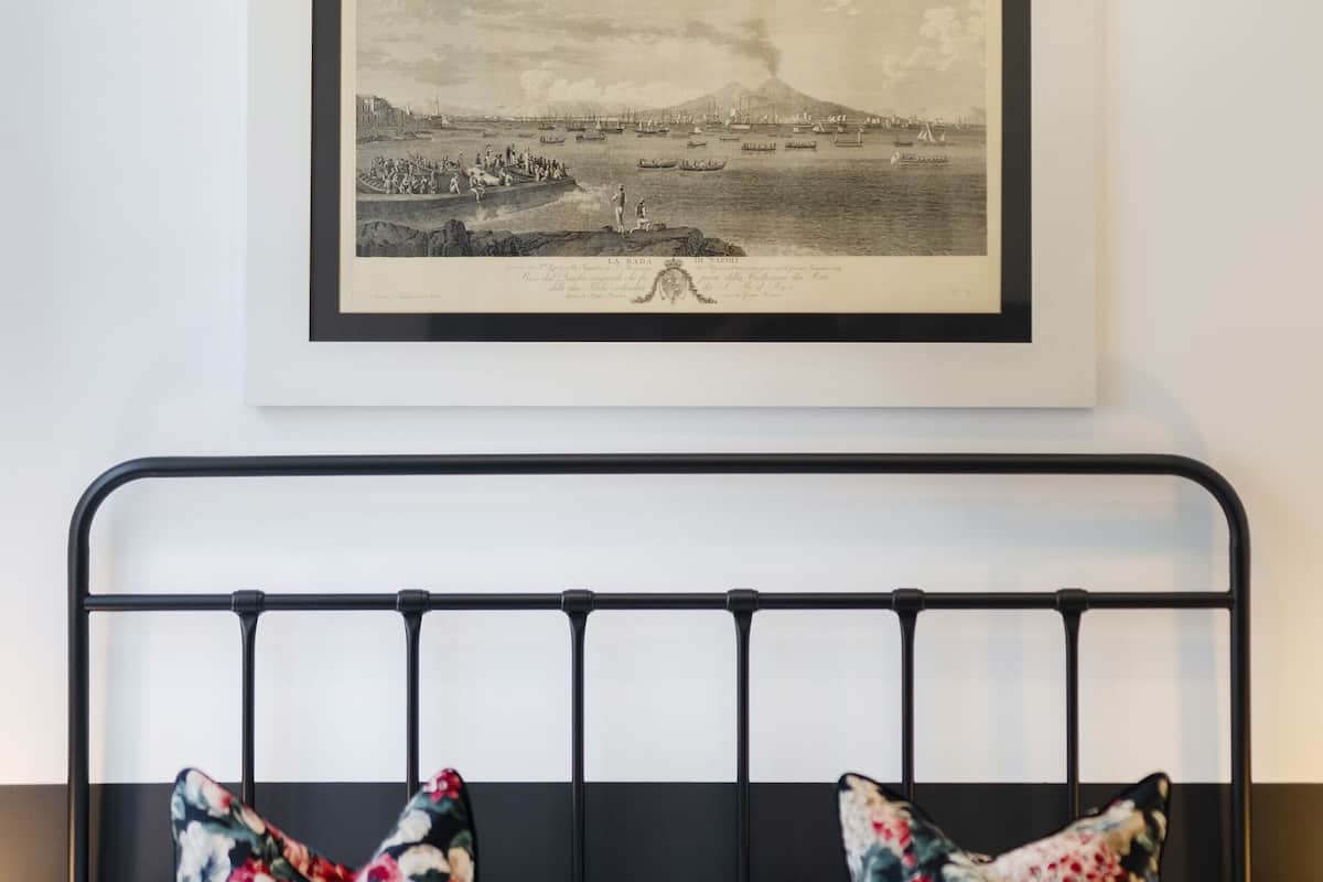 A neatly made bed with a black metal frame, two floral pillows, and a framed black and white landscape picture above it.