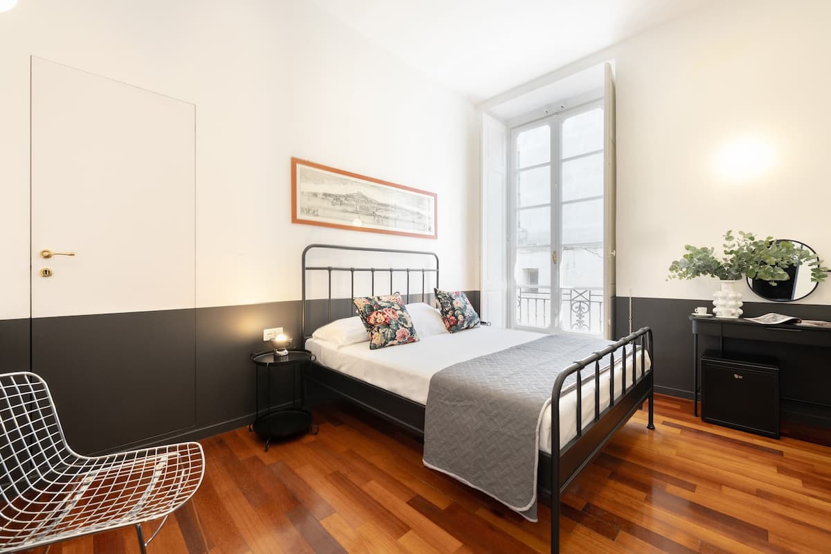 A bedroom with a bed, a chair, a bedside table, a nightstand, a window, a door, and a small cabinet.