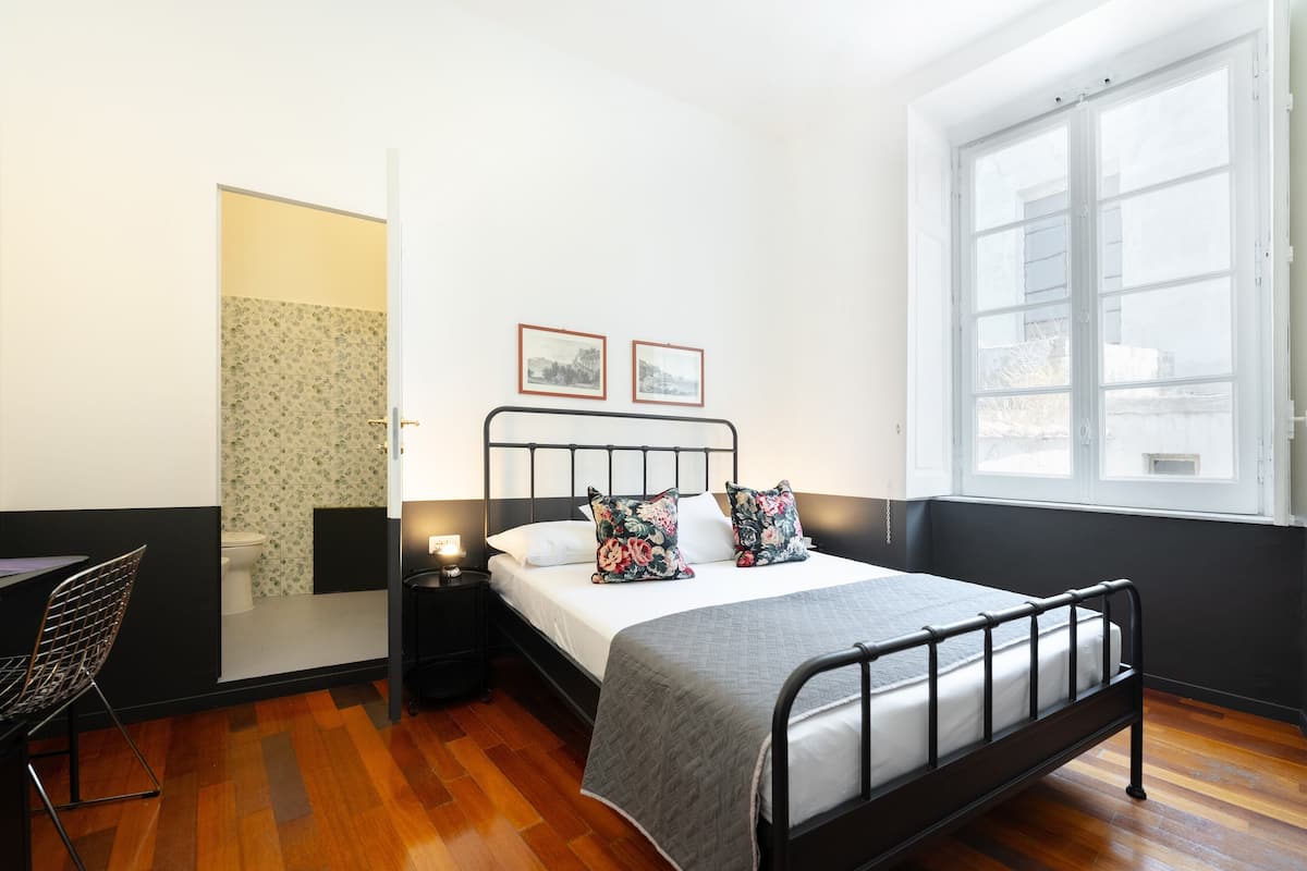 A bedroom with a bed, a chair, a nightstand, a window, and a door leading to a bathroom.