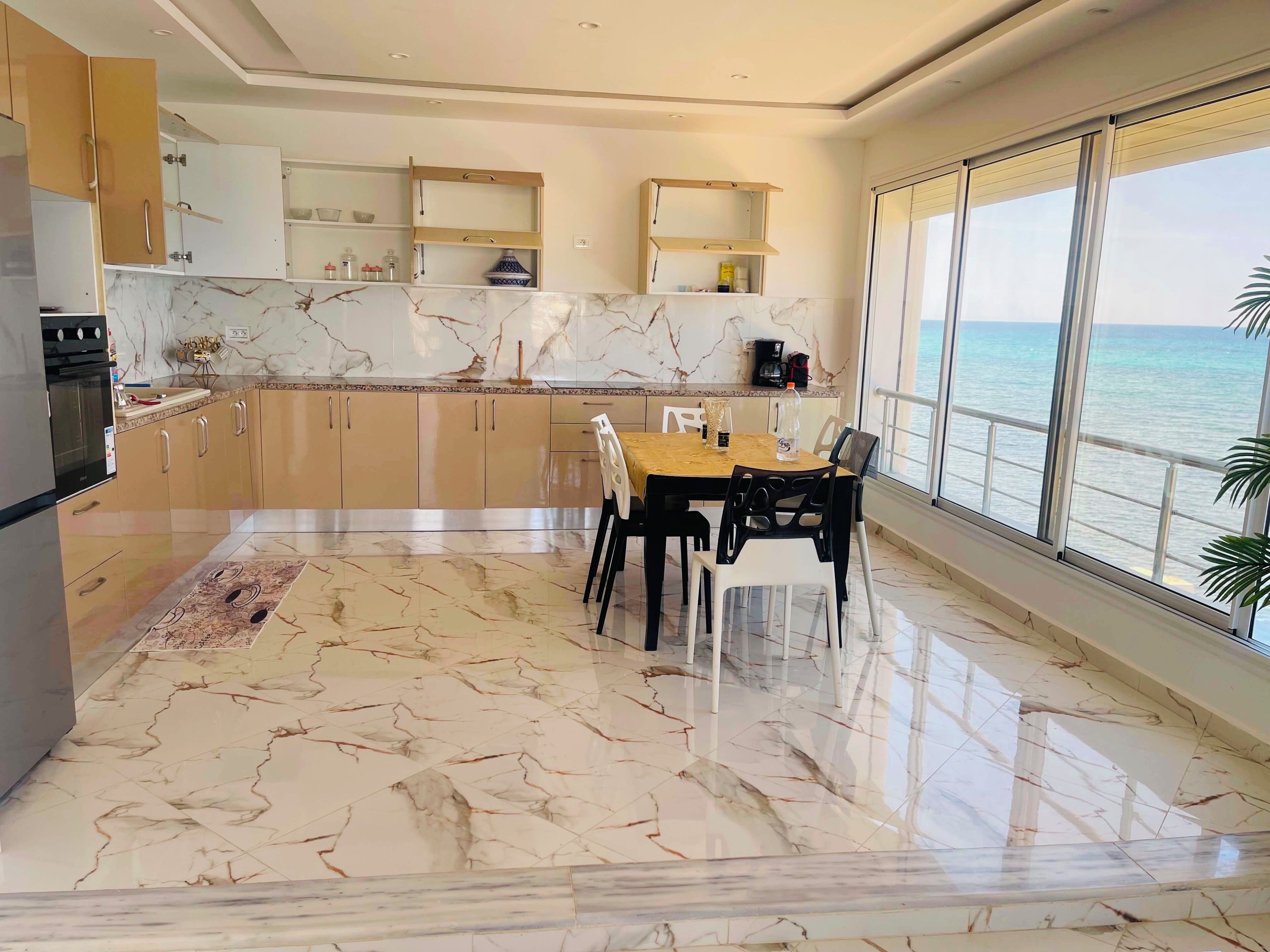 Una cocina moderna con suelo de mármol, una zona de comedor con mesa y sillas, y un amplio ventanal con vistas al mar.