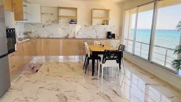 Una cocina moderna con suelo de mármol, una zona de comedor con mesa y sillas, y un amplio ventanal con vistas al mar.