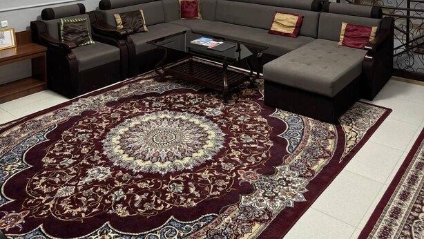 A modern living room with a sectional sofa, a glass coffee table, and a large patterned rug. A staircase with a wrought iron railing is visible in the background.