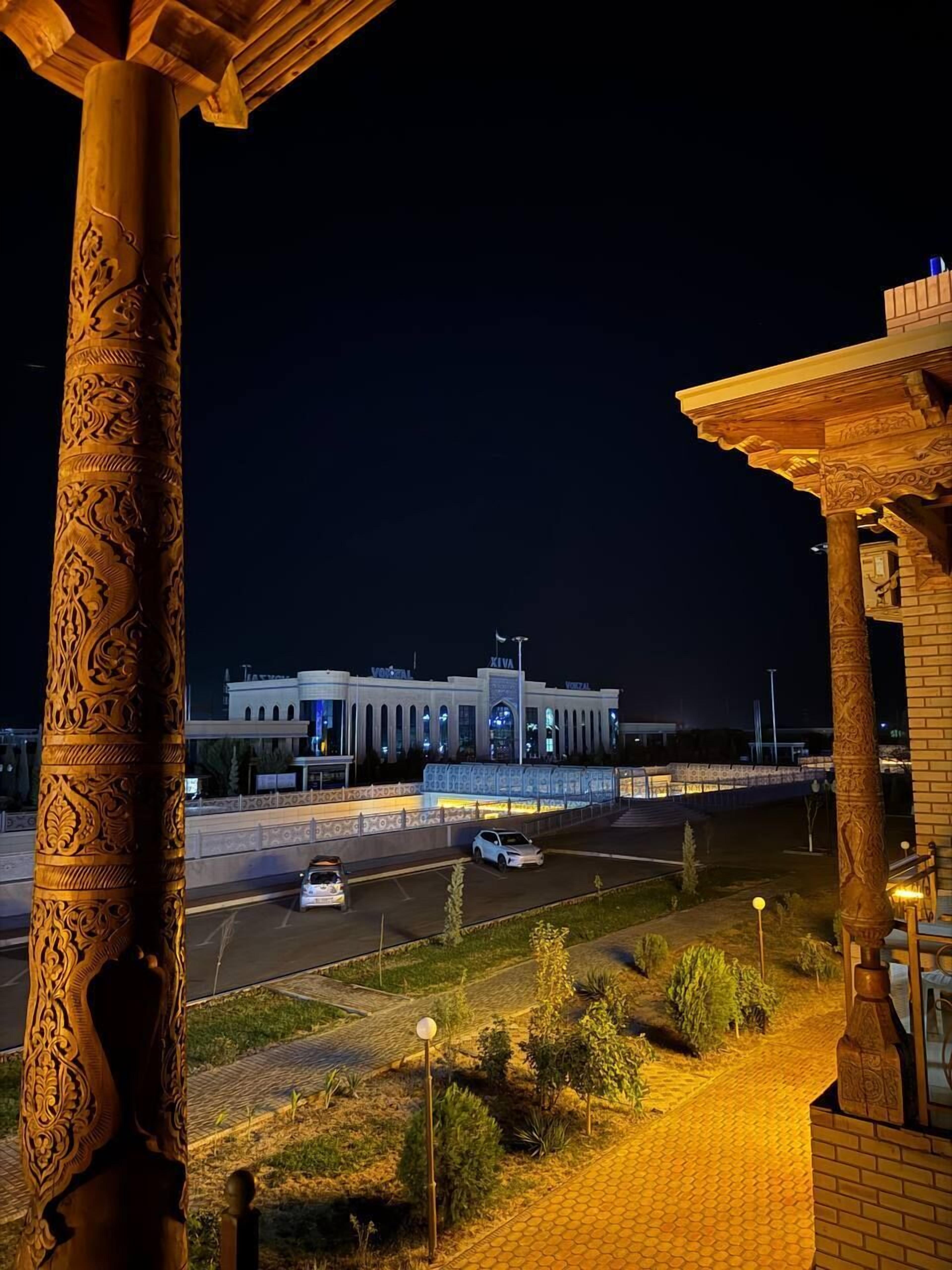 Una vista nocturna de un edificio con columnas de madera intrincadas y un camino pavimentado que conduce a un estacionamiento.