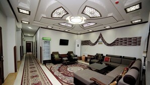 A spacious living room with a large sofa, a coffee table, and a patterned rug. There is a television mounted on the wall and a bookshelf in the corner.