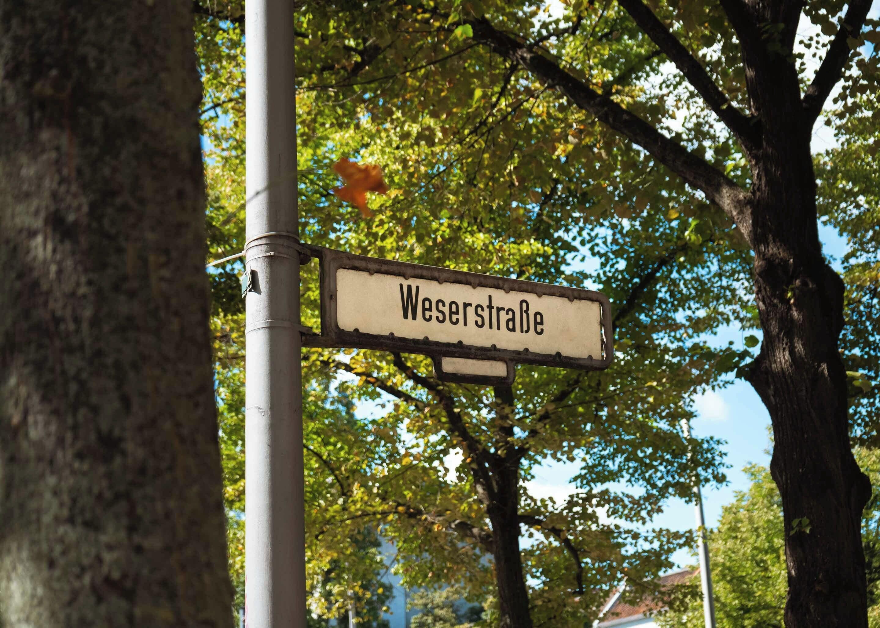A street sign reading "Weserstraße" mounted on a pole with trees in the background.