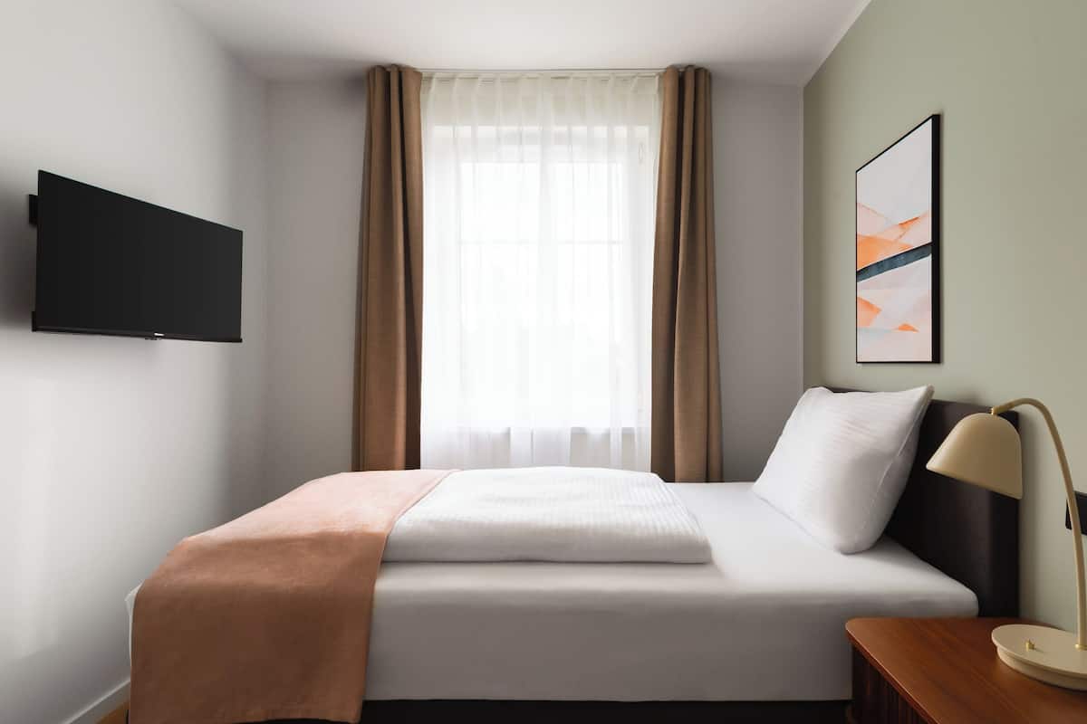 Habitación de hotel con cama, televisión montada en la pared, ventana con cortinas y un cuadro en la pared.