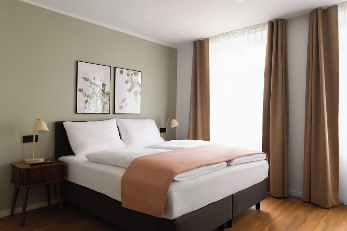 Una cama bien hecha con un edredón marrón, dos lámparas de noche y dos láminas enmarcadas con motivos botánicos en la pared.
