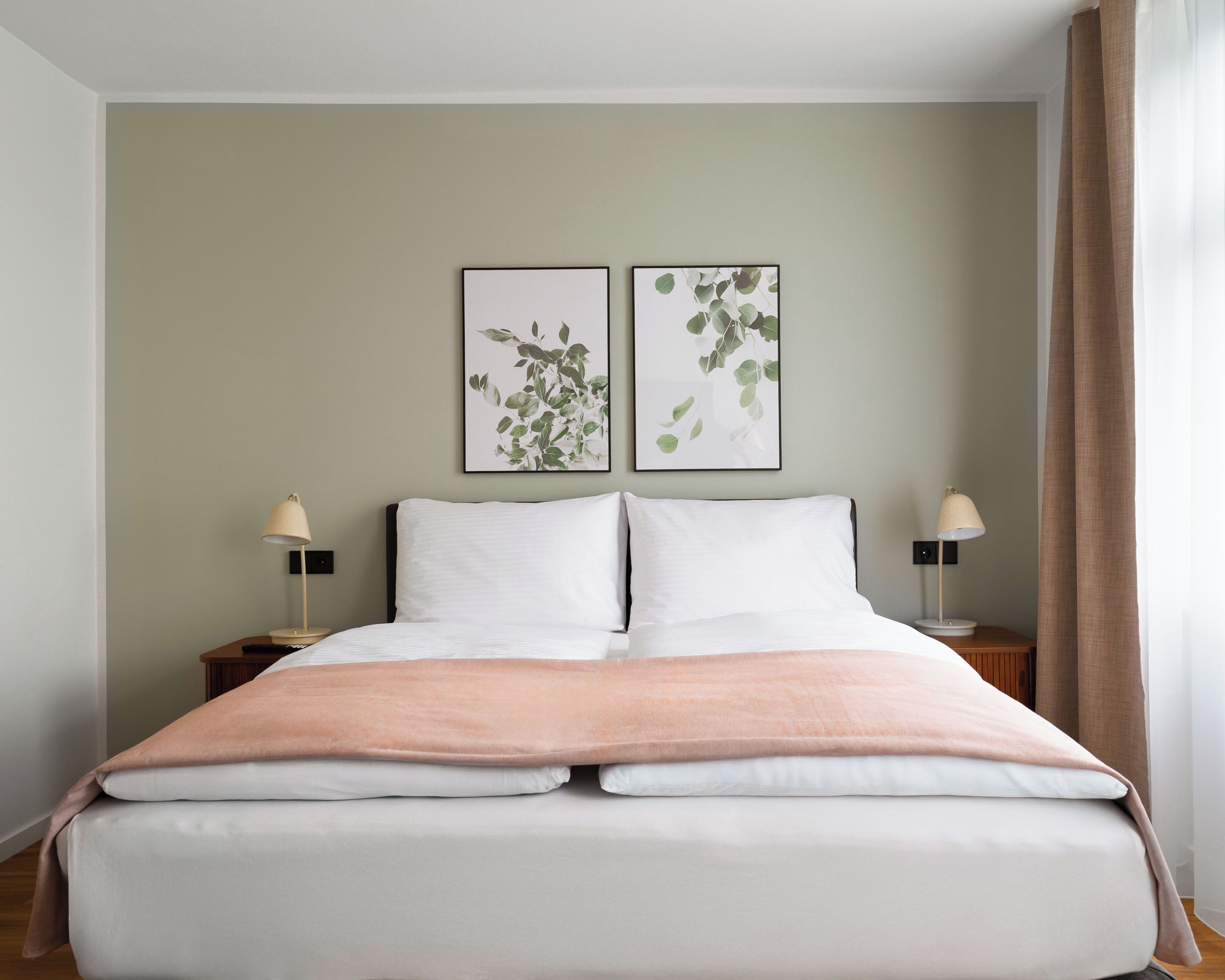 Un lit bien fait, avec une literie blanche et pêche, deux tables de chevet avec des lampes, et deux reproductions botaniques encadrées accrochées au mur.