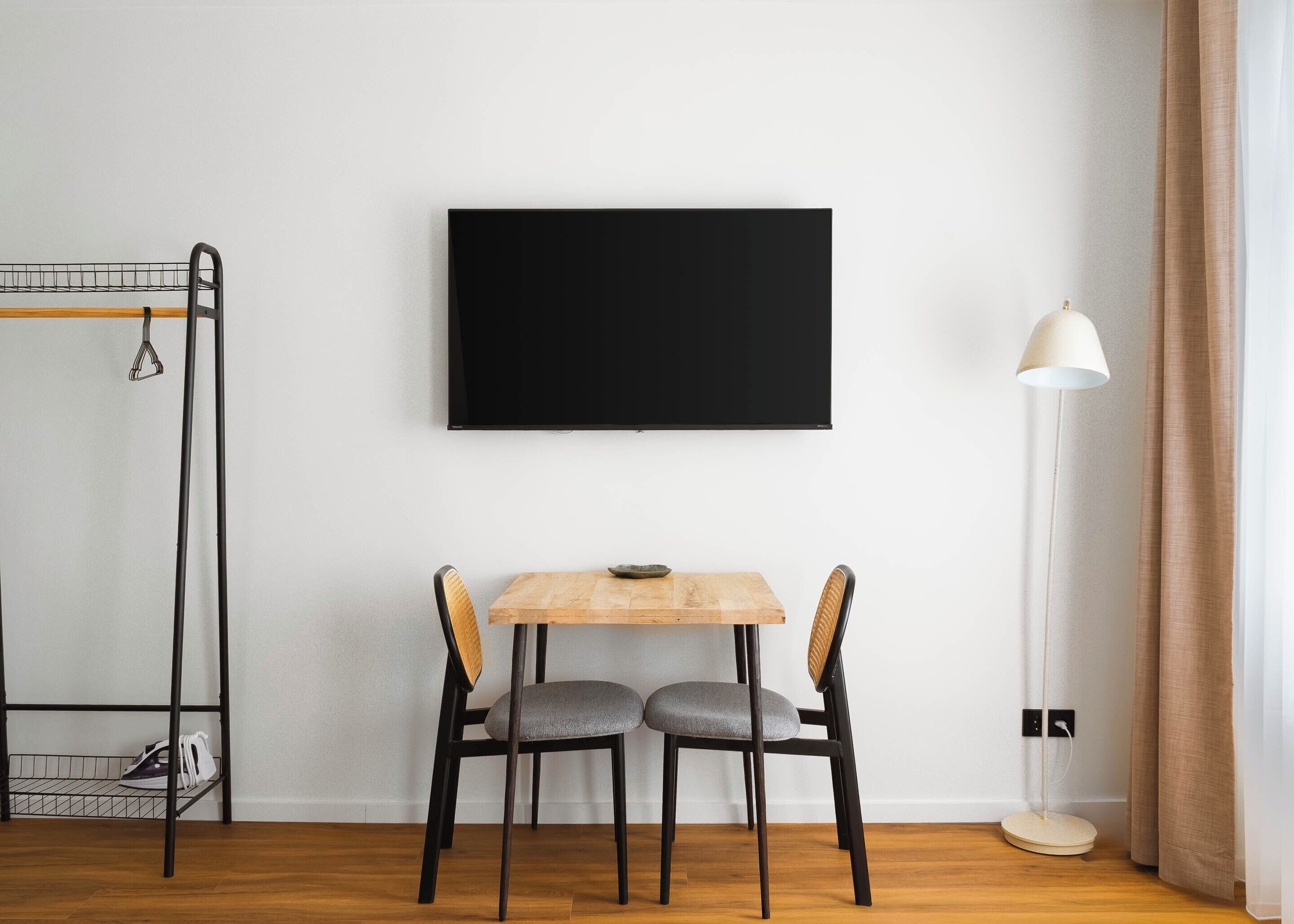 Habitació amb taula de fusta, dues cadires, un televisor negre a la paret, una làmpada de peu i un prestatge per sabates.