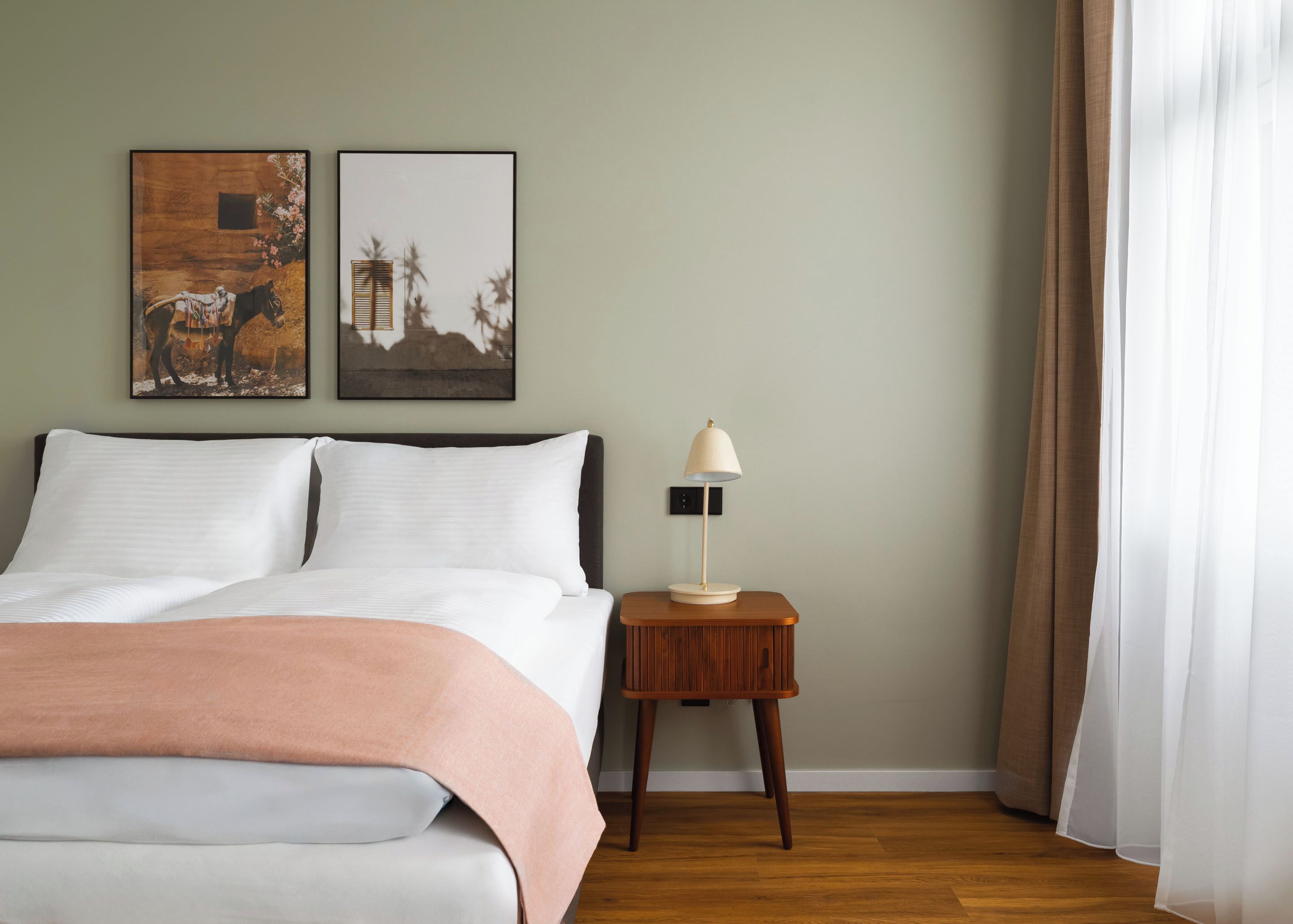 Un lit bien fait, recouvert d’une couverture blanche et pêche, une lampe de chevet et deux tableaux encadrés accrochés au mur.