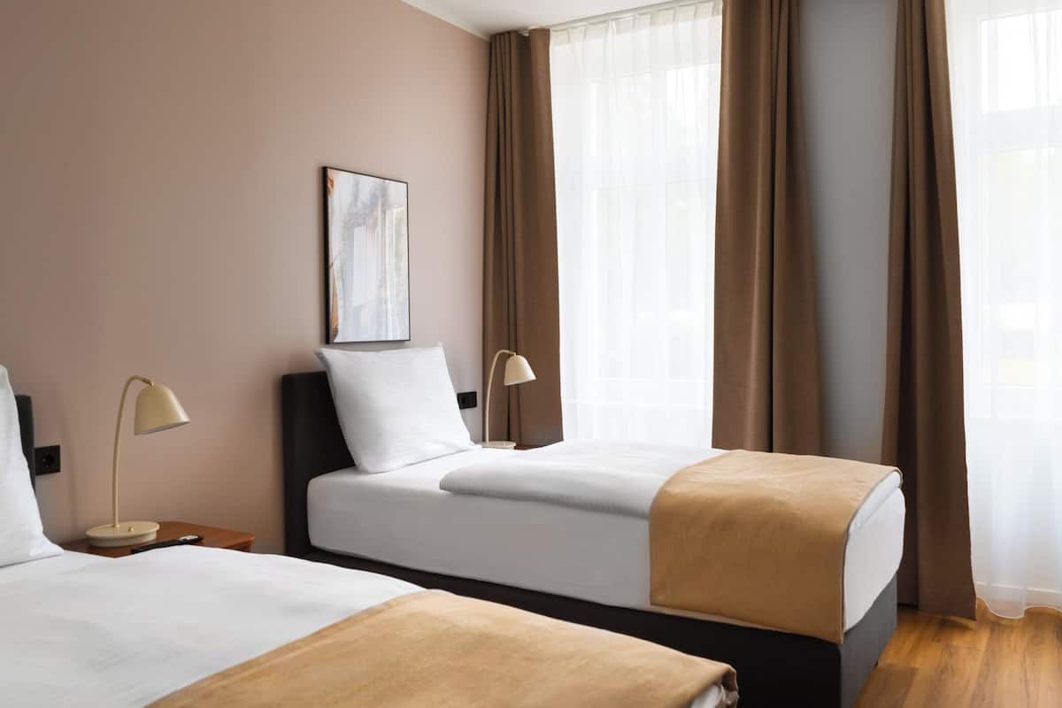 Habitación de hotel con dos camas, una mesita de noche con lámpara, un cuadro en la pared y amplias ventanas con cortinas.