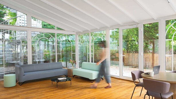 Uma sala de estar moderna com um sofá cinzento, uma mesa de centro e uma área de jantar com cadeiras e uma mesa. Grandes janelas oferecem vista para o verde lá fora.